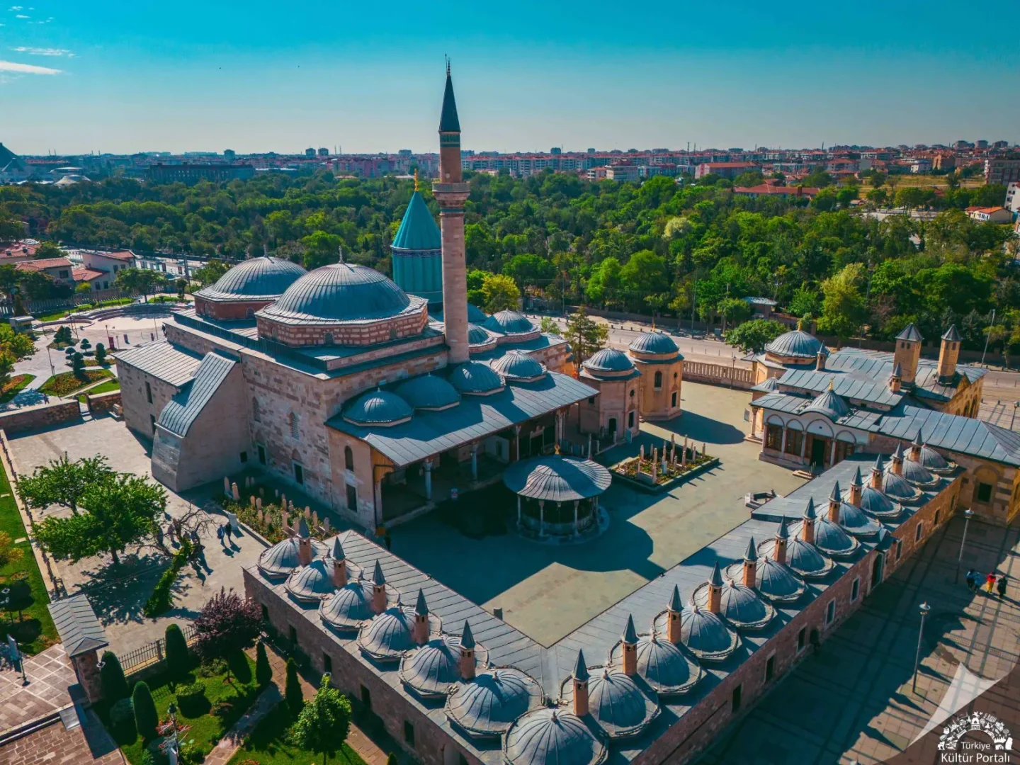 Konya Mevlana Müzesi'nin havadan çekilmiş bu geniş görüntüsünde, ikonik yeşil kubbesi ve ince minaresiyle birlikte, gri kubbeleri ve taş binalarıyla çevrili avlusu, yeşil ağaçlar ve şehir manzarasıyla bir bütün olarak güneşli bir günde göz kamaştırıyor.