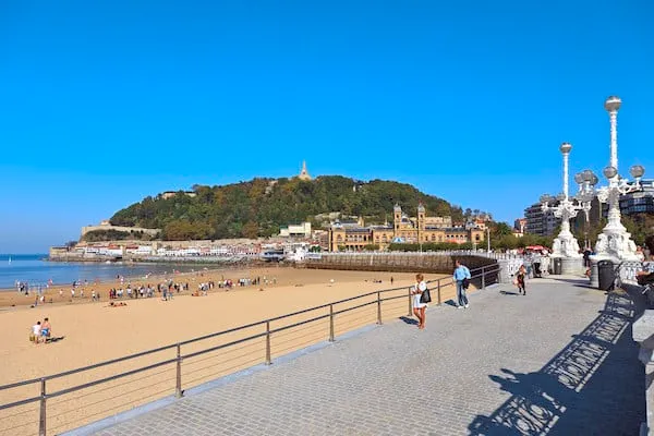 İspanya'nın San Sebastián şehrindeki, hilal şeklindeki La Concha Koyu'nun huzurlu manzarasını gösteren bu fotoğrafta, altın rengi kumsalın etrafını saran turkuaz sular, körfezin ortasındaki Santa Clara Adası ve sahil boyunca sıralanmış tarihi binalar güneşli bir havada parlıyor.