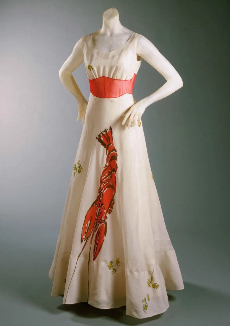 Elsa Schiaparelli'nin 1937'de Salvador Dalí ile iş birliği yaparak tasarladığı, ön tarafında çarpıcı kırmızı bir ıstakoz deseni bulunan, saten veya ipek gibi lüks bir kumaştan yapılmış, beyaz ve zarif Istakoz Elbisesi, sürrealizm akımının moda dünyasındaki etkileyici bir yansıması olarak zamana meydan okuyan ikonik bir eser.