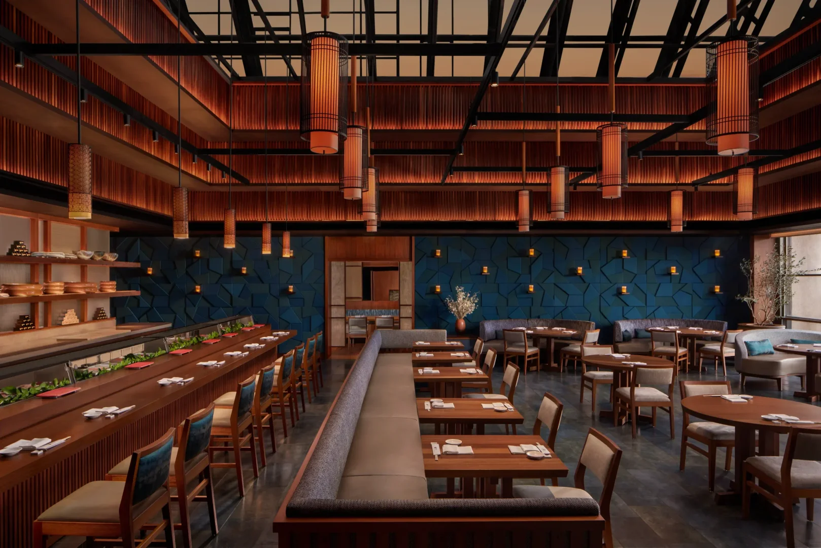 Nobu İstanbul restoranının modern ve şık iç tasarımı, uzun ahşap bir suşi barı, koyu teal rengi geometrik desenli duvarları, ahşap masa ve sandalyelerin yanı sıra tavandan sarkan sıcak ışıklı silindirik lambalarıyla göz alıcı bir atmosfer sunuyor.