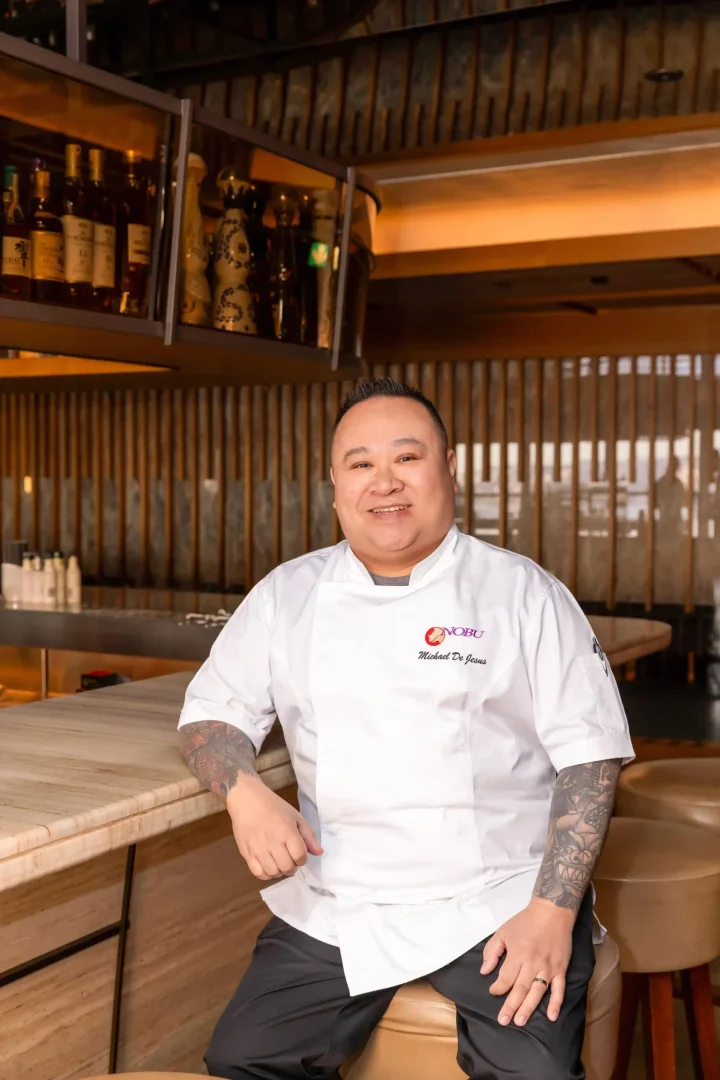 Nobu İstanbul'un Baş Aşçısı Michael De Jesus, şık ve modern restoranın ahşap detaylarla bezeli barında, üzerinde Nobu logosu ve adının yazılı olduğu beyaz şef ceketini ve dövmeli kollarını sergileyerek kameraya içten bir gülümsemeyle bakıyor.