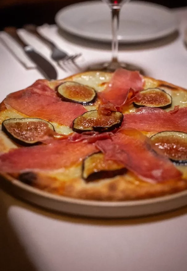 Pizzeria Pera'da servis edilen, erimiş beyaz peynir, ortadan ikiye kesilmiş koyu renkli incir dilimleri ve ince pembe füme jambonlarla bezenmiş, arka planda kadeh ve çatal bıçak takımının görüldüğü iştah açıcı bir pizza.