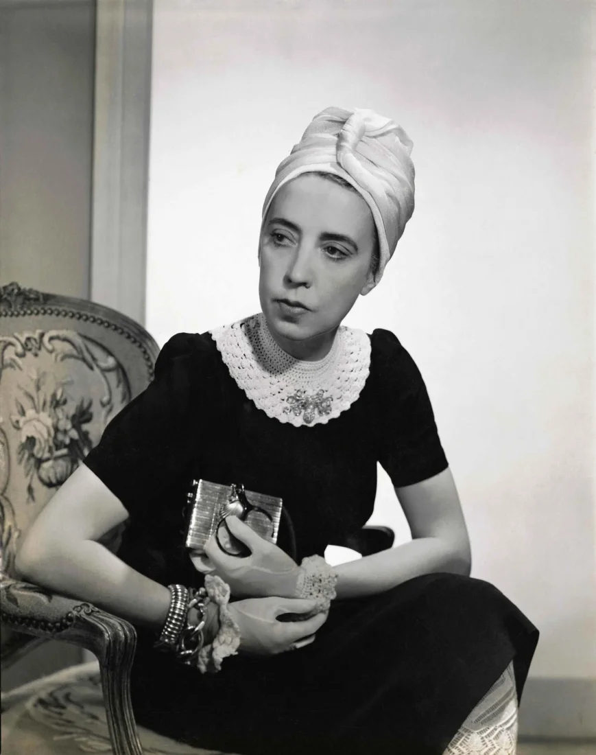 Elsa Schiaparelli, 1940'ların özgün ve yenilikçi moda dünyasına yön veren ünlü bir tasarımcı olarak, şık kıyafeti ve karizmatik duruşuyla dikkat çeken, sofistike bir portrede resmedilmiş.