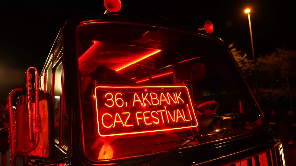 Akbank Caz Festivali logosu ve adını taşıyan koyu renkli, şık bir minibüs, kalabalık bir şehir caddesinde beklerken, festivalin erişilebilirliğini ve cazın dinamik ruhunu yansıtan mobil bir deneyim sunarak müzikseverleri etkinliklere davet ediyor.