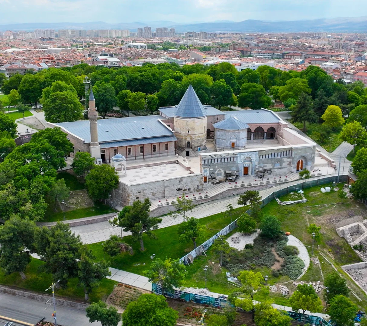 Konya'daki tarihi Alaaddin Camii ve bitişiğindeki sivri konik çatılı Sultanlar Türbesi'nin yemyeşil ağaçlarla çevrili bir park içinde kuşbakışı görüntüsü, arka planda şehrin binalarıyla uzanan bir şehir manzarası ve dağ silüetiyle bütünleşiyor.