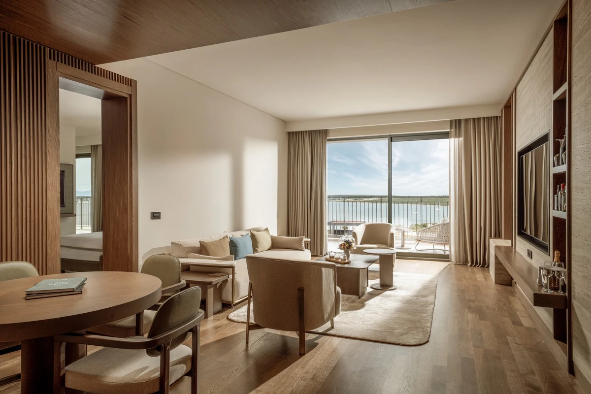 Anda Barut Collection'a ait modern tasarımlı, ahşap detaylarla zenginleştirilmiş geniş bir Başkanlık Süiti oturma alanı; solda yemek masası, ortada konforlu koltuk takımı ve kahve tabağındaki ikramlarla, büyük cam kapılarından uzanan ferah deniz manzaralı balkon ile lüks bir konaklama deneyimi sunuyor.