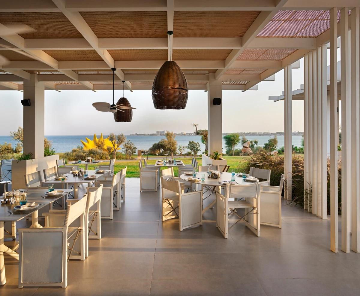 Anda Barut Collection'ın Balık Kampı Alakart Restoranı'nın, denize nazır yemyeşil bir bahçeye bakan, modern beyaz mobilyalar, hasır avizeler ve tavan vantilatörüyle donatılmış, geniş ve huzurlu açık hava yemek alanı.