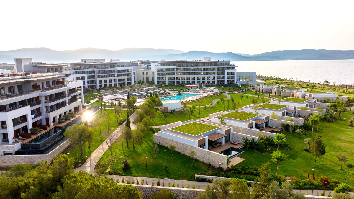 Bodrum'daki Anda Barut Collection'ın tepeden çekilmiş bu genel görünümünde, yemyeşil çatıları olan modern taş villalar, özel havuzlu terasları olan çok katlı otel binaları, büyük bir ana havuz alanı ve pırıl pırıl denize uzanan geniş peyzajlı bahçeler göze çarpıyor.