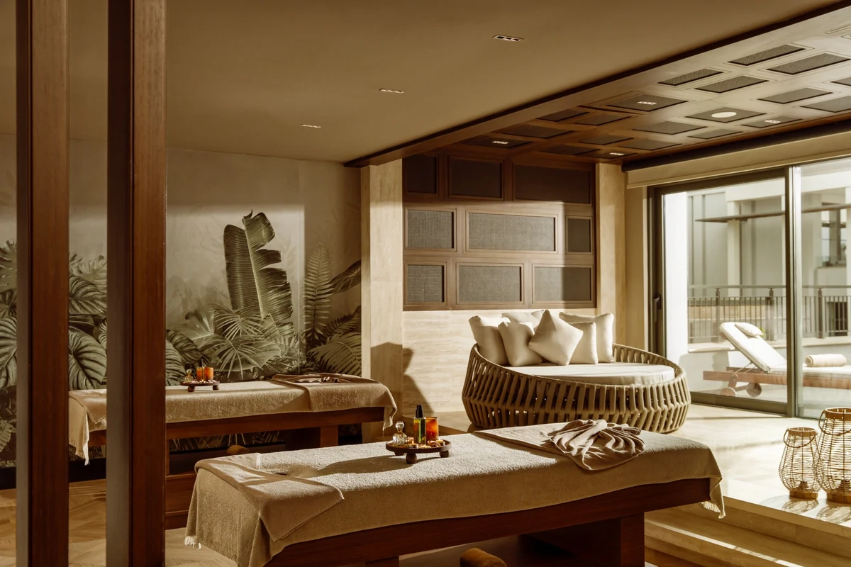 Bu lüks ve huzurlu spa odası, bej havlularla örtülü iki ahşap masaj yatağı, konforlu yuvarlak dinlenme koltuğu, tropik yapraklı duvar desenleri ve dışarıdaki şezlonglu teras manzarasıyla misafirlere keyifli bir dinlenme deneyimi vadediyor.