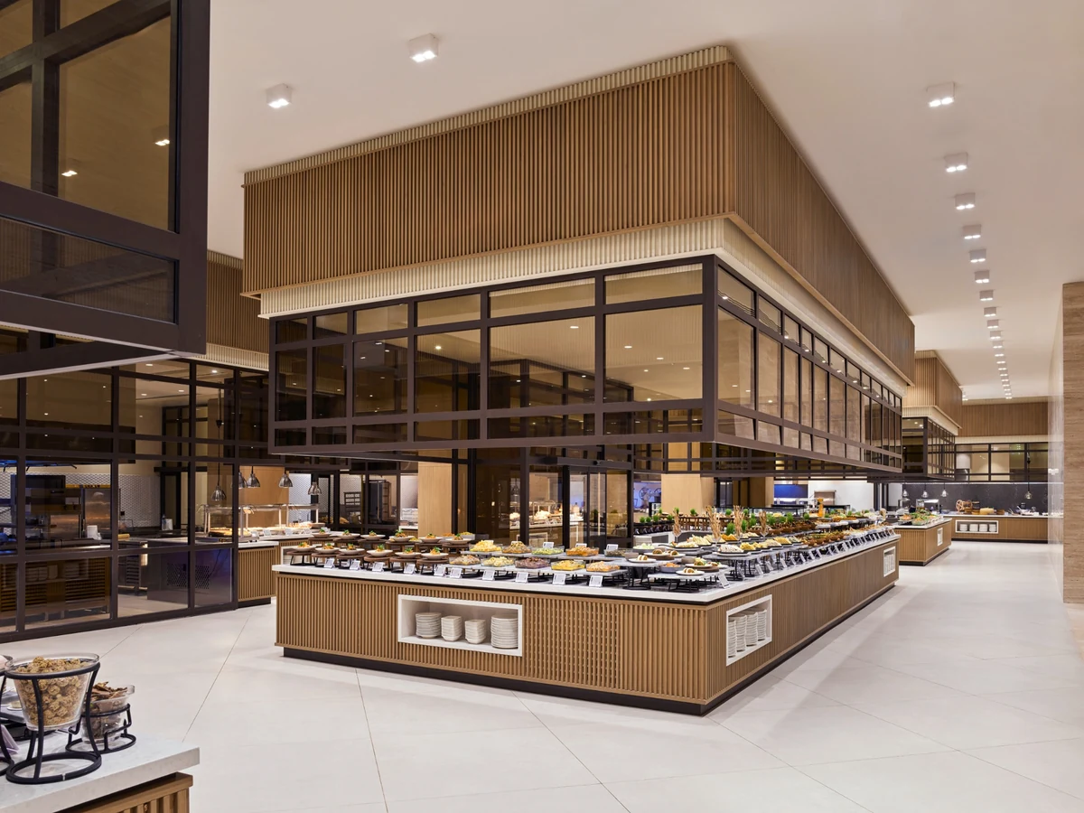 Anda Barut Collection'ın Zestful Bazaar ana restoranında, çok sayıda lezzetli yiyecekle donatılmış uzun, modern ahşap büfe ve cam bölmelerle ayrılmış mutfak istasyonlarının bulunduğu, misafirler için davetkar ve ferah bir yemek alanı.