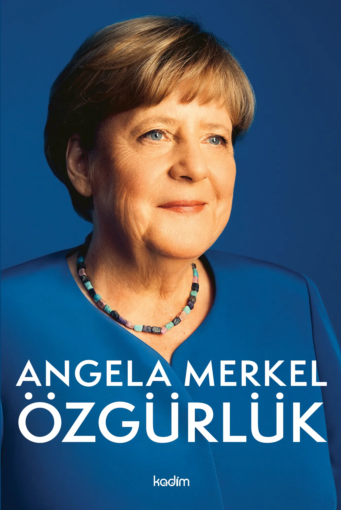 Angela Merkel, açık renk bir blazer ceket giymiş ve ellerini önünde kavuşturmuş şekilde dururken, gülümseyerek ileriye bakıyor ve liderliğiyle özgürlük kavramına yaptığı katkıları yansıtıyor.