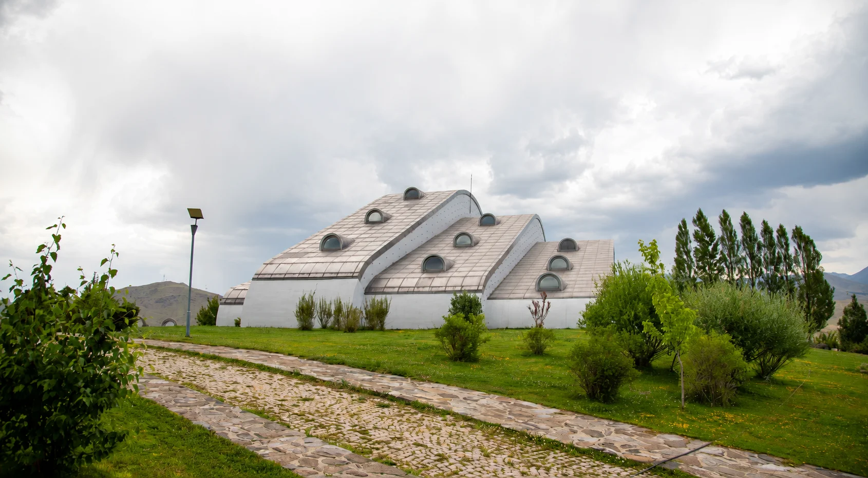 Baksı Müzesi'nin geniş açık hava sergi alanında, modern ve geometrik formlara sahip, parlak metalden yapılmış heykelimsi bir çağdaş sanat eseri, arka planda uzanan mimari yapı ve uzaklardaki doğal manzarayla bütünleşirken, bir ziyaretçi bu etkileyici eseri dikkatle inceliyor.