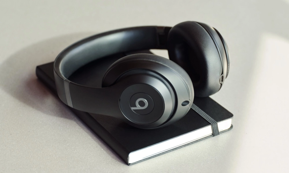 Açık gri bir zeminde, sol kulaklığında belirgin Beats logosu olan mat siyah Beats Studio Pro kablosuz kulaklık, kapalı siyah bir not defterinin üzerinde şık bir şekilde duruyor ve modern, minimalist bir estetik sunuyor.