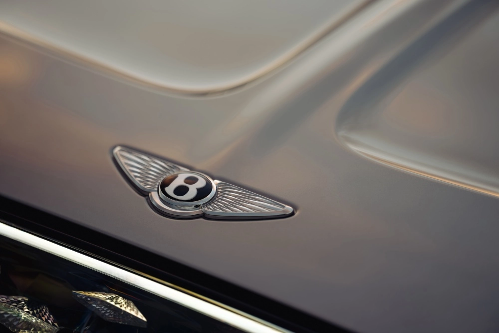 Yeşilliklerle çevrili bir dış mekanda park halinde duran koyu renkli, lüks Bentley SUV'nin ön-yan görünümü, yazıda bahsedilen 888 beygir gücünde ve 600 km menzilli Bentley Torcal elektrikli SUV'nin aerodinamik ve güçlü tasarımını vurguluyor.