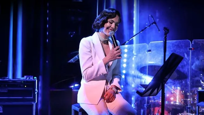 Mavi ve mor sahne ışıkları altında, beyaz şık bir takım elbise ve inci kolye takan, kısa dalgalı saçlı Bergüzar Korel, gülümseyerek oturup elinde mikrofon tutarak canlı bir konser performansı sergiliyor.
