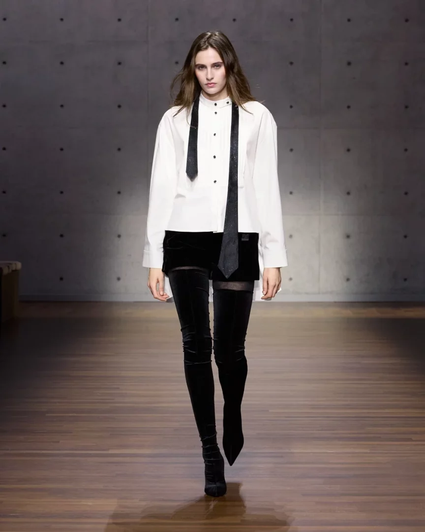 Bir podyumda yürüyen, beyaz uzun kollu gömlek giymiş, boynunda simli siyah kravat sarkıtan, siyah kadife şort ve diz üstü çizmelerle modern bir tarz sergileyen model, Armani beyaz gömlek rehberi içeriğine uygun güçlü bir duruş sergiliyor.