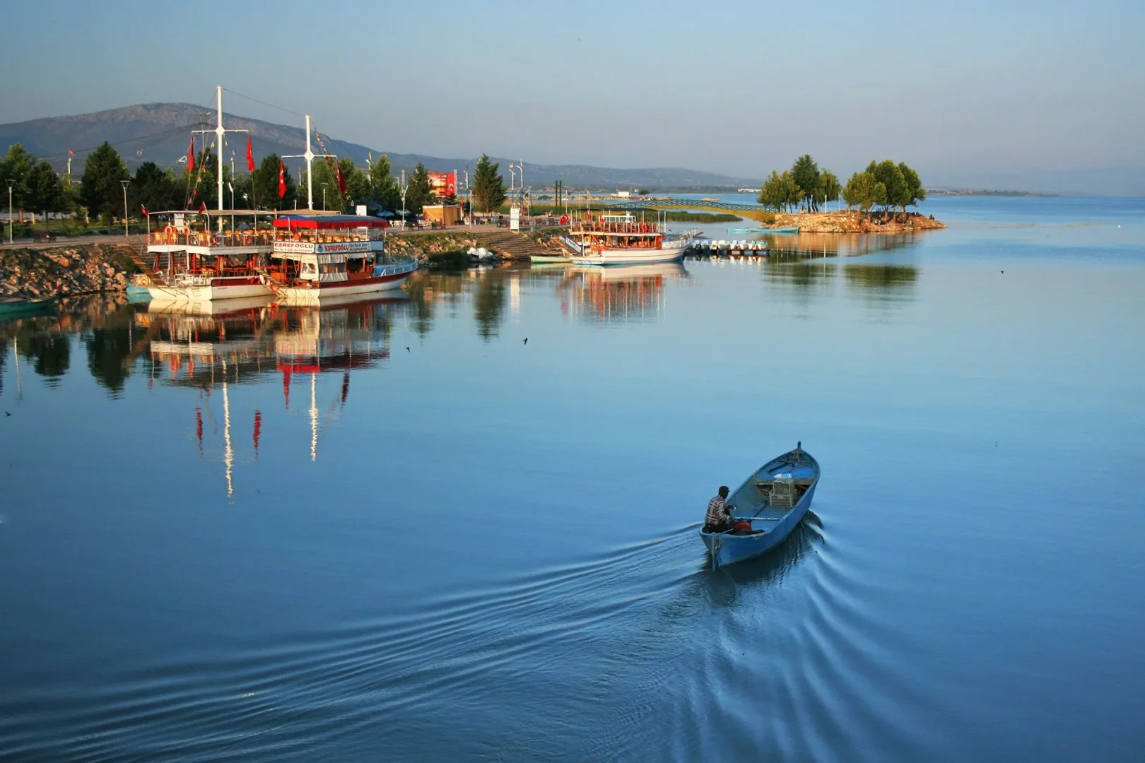 Beyşehir Gölü'nün huzur veren sularında bir kişinin motorlu teknesiyle ilerleyerek geride iz bıraktığı, sol tarafta Türk bayraklı büyük gezi teknelerinin demirli olduğu, yemyeşil ağaçlarla bezeli kıyı şeridinin, küçük adacığın ve uzakta yükselen dağların berrak yansımalarının görüldüğü geniş açı bir manzara.