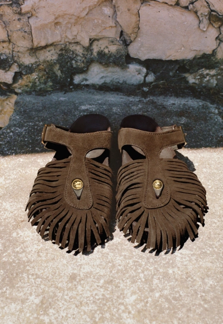 Koyu kahverengi süet benzeri dokulu, ön kısımları püsküllü ve ortasında altın rengi yuvarlak detaylara sahip bir çift Birkenstock Etro Gizeh sandalet, eskitilmiş taş duvarın önündeki açık renkli dokulu bir zemin üzerinde yakın çekim olarak sergileniyor.