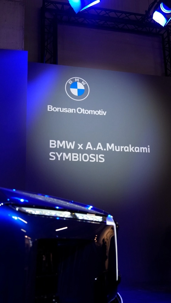 BMW 7 Serisi'nin lüks ve modern iç mekanında, ön yolcu tarafı konsol ve kapı paneli detaylıca gösteriliyor; dijital ekranın yanındaki parlak süsleme ve kapı boyunca uzanan özel desenli şerit, Murakami Symbiosis işbirliğinin özgün estetiğini yansıtıyor.