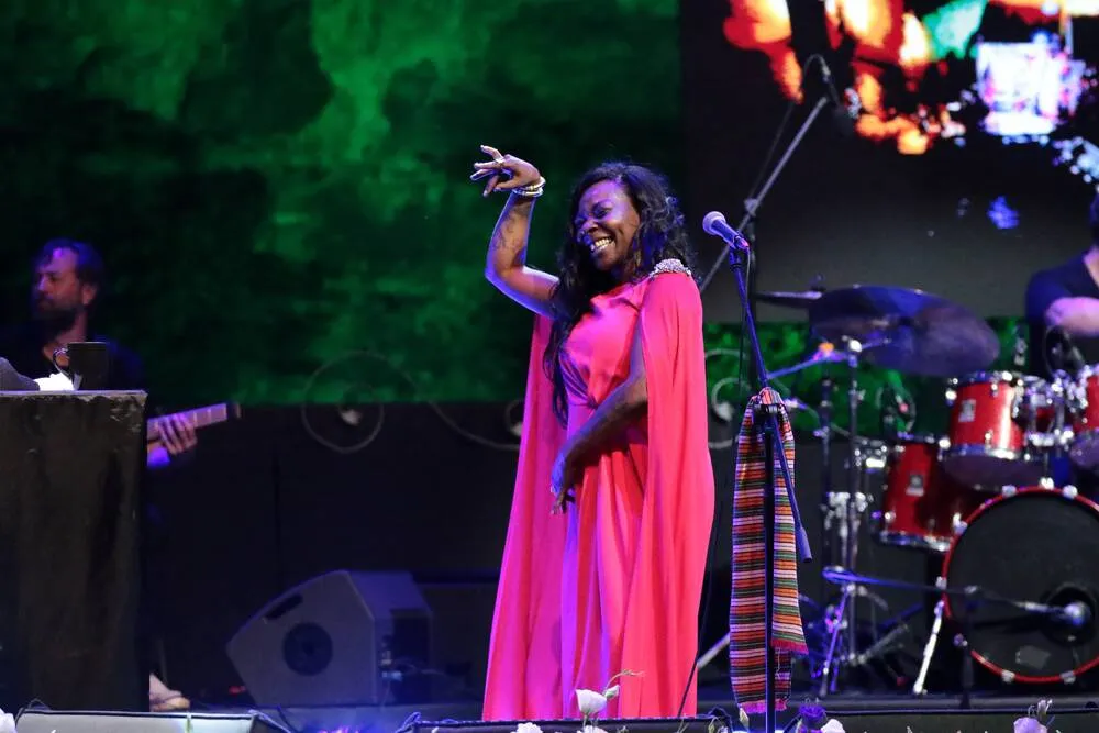 Buika, koyu renk kıvırcık saçları, açık renk bluzu ve büyük güneş gözlükleriyle, önündeki bir mikrofonla açık havada bir masada otururken bir caz festivali röportajı sırasında düşünceli bir ifadeyle dinleyiciye veya muhabire bakıyor.