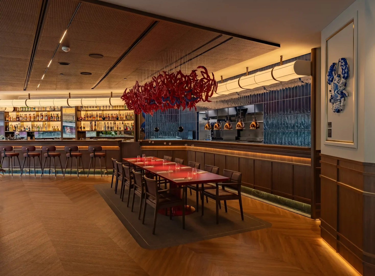 Modern ve şık bir restoranın iç mekanında, uzun bir bara dizili içki şişeleri, on iki kişilik kırmızı bir yemek masasının üzerindeki kırmızı ıstakoz figürlü avize ve duvardaki büyük mavi beyaz ıstakoz sanatı dikkat çekmekte olup, mekana özel bir atmosfer katmaktadır.