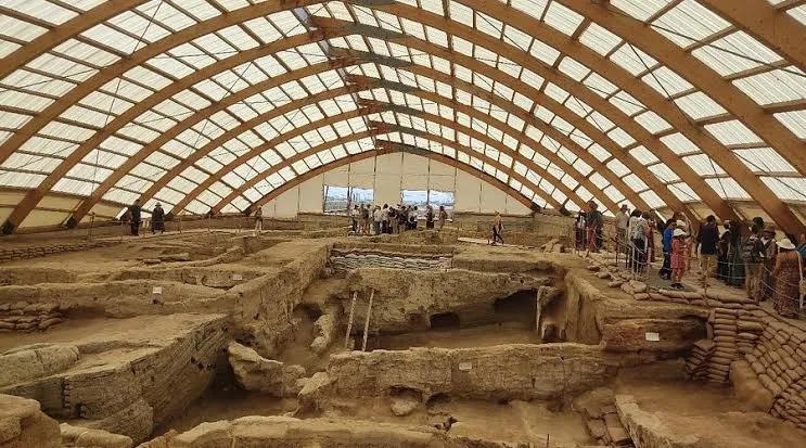 Büyük, kemerli, ahşap ve yarı şeffaf panelli bir çatı, Çatalhöyük arkeolojik kazı alanını kaplamakta; burada çok sayıda antik kerpiç yapı ve kazı katmanları belirgin bir şekilde görülürken, ziyaretçi grupları yükseltilmiş yollardan bu Neolitik yerleşimi incelemektedir.