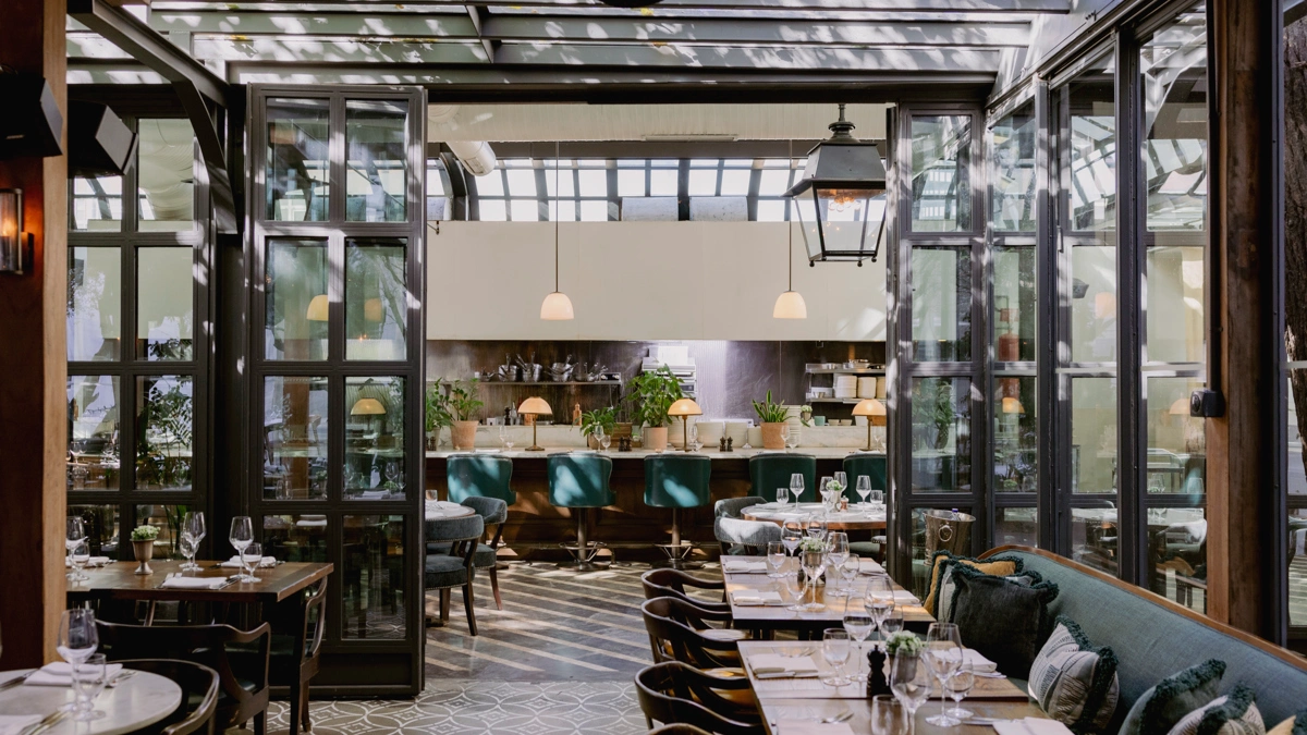 Cecconi's restoranının zarif iç mekanı, beyaz masa örtülü yuvarlak masaları, koyu yeşil döşemeli şık sandalyeleri ve geniş pencerelerden içeri süzülen bol doğal ışık ile davetkar bir yemek atmosferi sunarak, yeşil bitkiler ve özenli dekorasyon detaylarıyla misafirlerine unutulmaz bir yeme içme deneyimi vaat ediyor.
