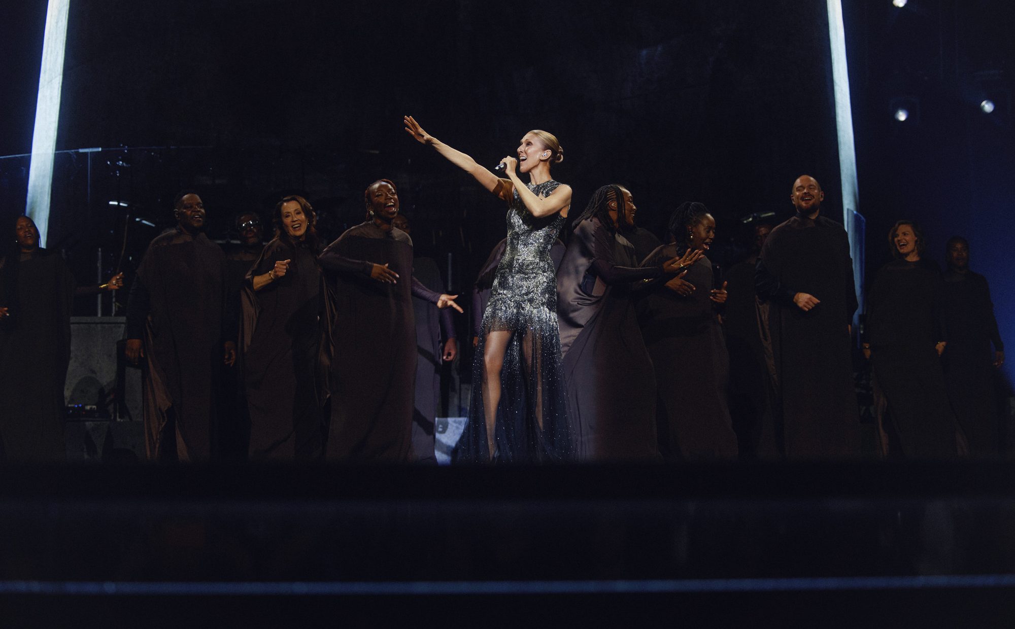 Celine Dion, parıltılı kısa elbisesiyle sahne ortasında mikrofon tutarak ve bir kolunu yukarı kaldırarak coşkulu bir şekilde şarkı söylerken, koyu renk uzun elbiseler giymiş çok sayıda koro üyesi de ona eşlik ederek karanlık sahneyi aydınlatıyor.