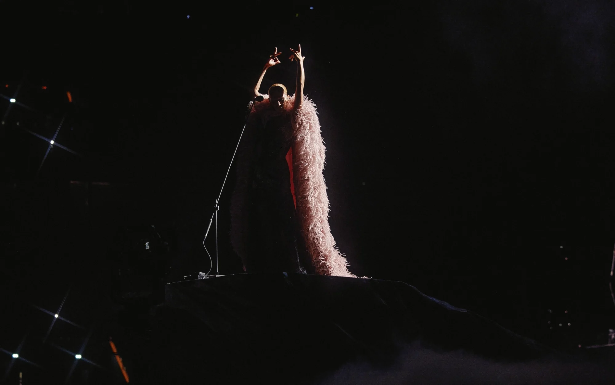 Celine Dion, karanlık sahne üzerinde spot ışıklarıyla aydınlatılmış, uzun, tüylü, açık renk kostümü içinde kollarını havaya kaldırmış, güçlü bir konser performansı anında izleyicilere hitap ediyor, solunda bir mikrofon standı dikkat çekiyor.