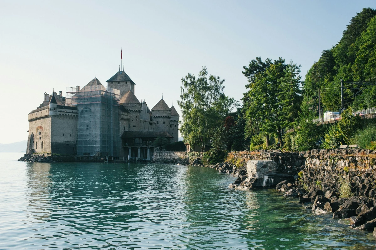 Cenevre gezilecek yerler arasında yer alan ve Leman Gölü'nün turkuaz sularında yükselen tarihi Chillon Şatosu, bir cephesinde iskelelerle restorasyon çalışmaları sürerken, sağında yemyeşil ağaçlarla kaplı kayalık bir kıyı şeridi ve dik yamaç manzarasıyla dikkat çekiyor.