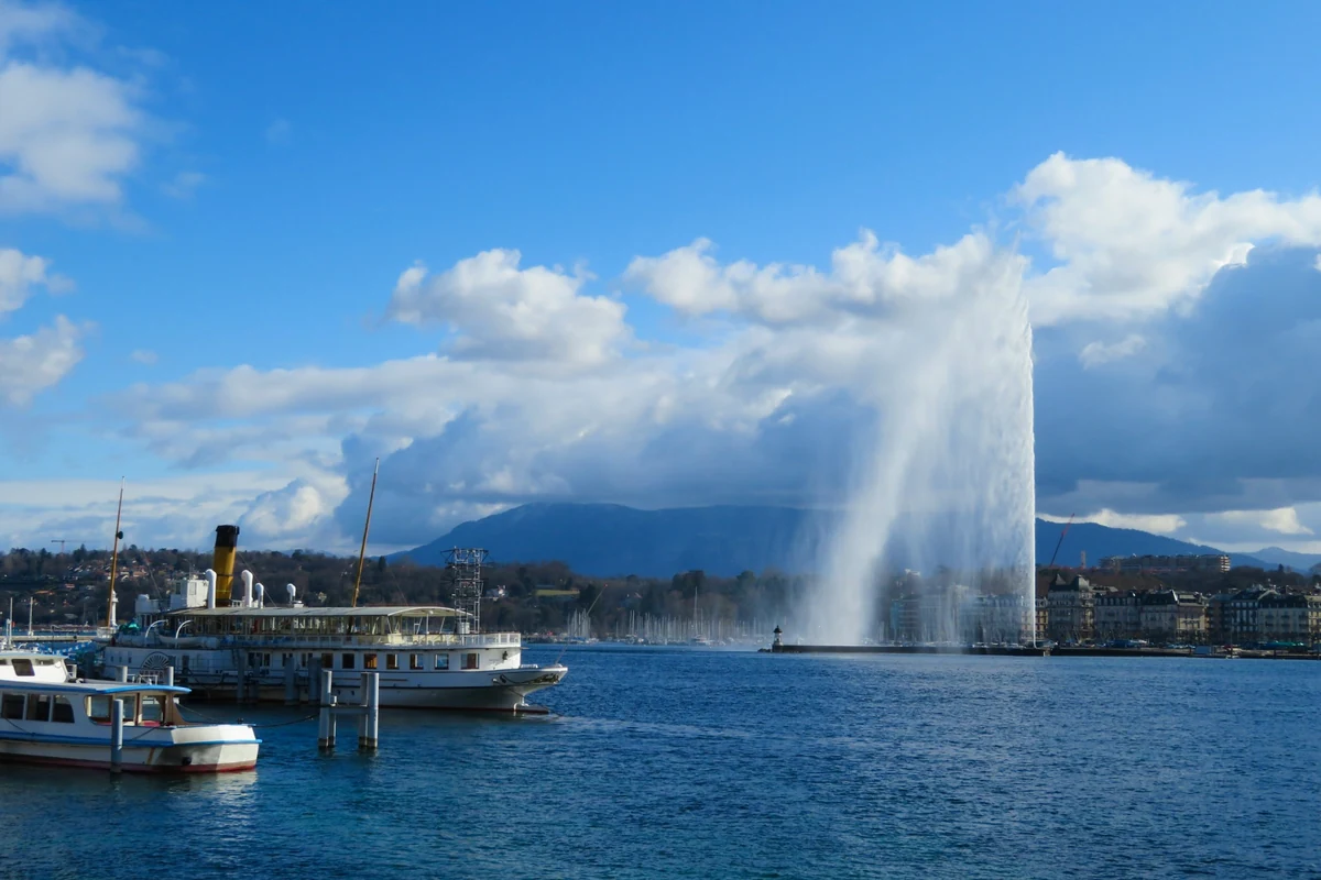 Cenevre'nin ünlü gezilecek yerlerinden Leman Gölü'nde, masmavi gökyüzü altında, meşhur Jet d'Eau fıskiyesinin ihtişamla göğe yükselen beyaz su sütunu, kıyıda demirli zarif bir vapur ve küçük bir tekne ile dağlarla çevrili şehir silüetiyle bütünleşiyor.