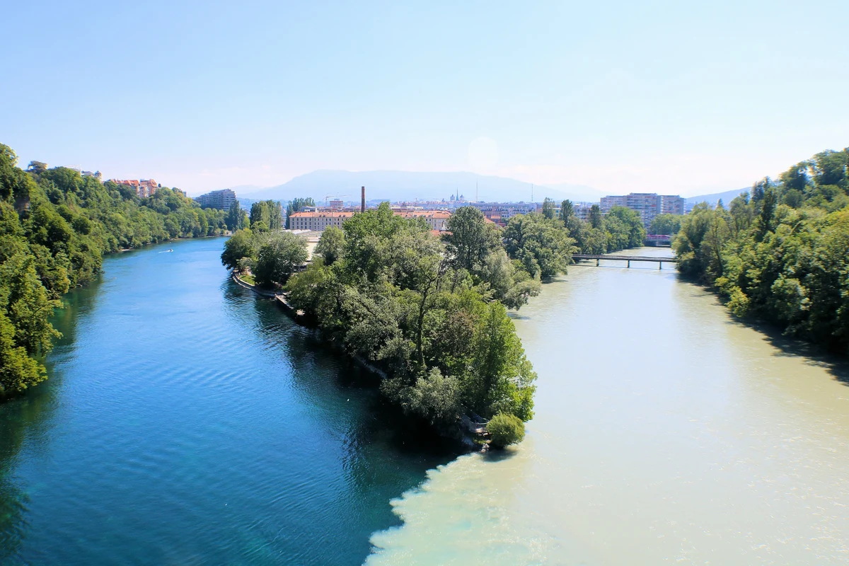 Cenevre'deki La Jonction bölgesinde Rhône ve Arve nehirlerinin berrak turkuaz ile çamurlu bej sularının ağaçlarla kaplı bir adanın etrafında birleştiği, şehir binaları ve dağların oluşturduğu arka planla büyüleyici bir manzara sunan doğal güzellik.