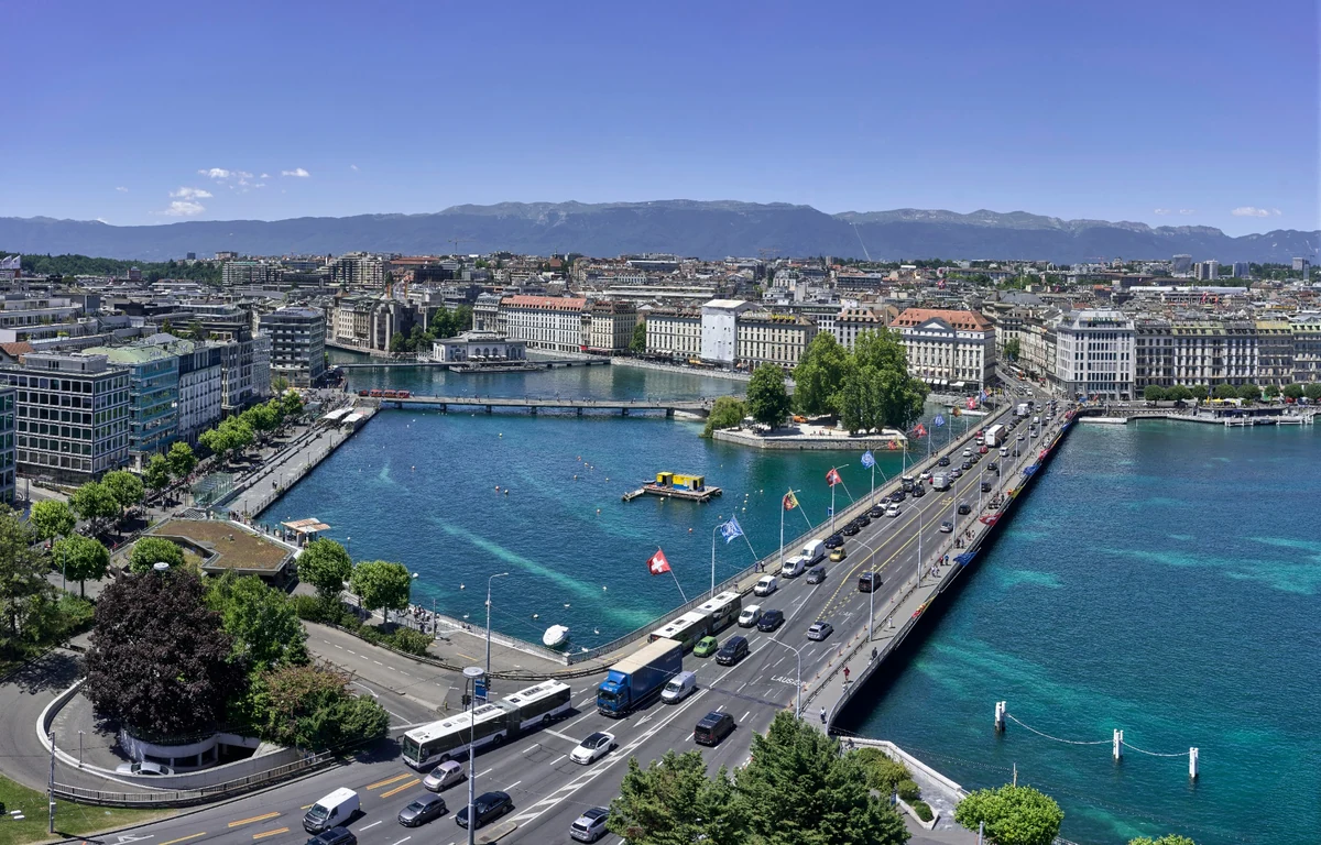Cenevre'nin hareketli şehir manzarası, masmavi göl suları üzerinde uzanan yoğun trafiğe sahip bir köprüyü ve arka planda yükselen dağlarla çevrili binaları gözler önüne seriyor, şehri gezilecek yerler listesine eklemek için cazip bir tablo çiziyor.