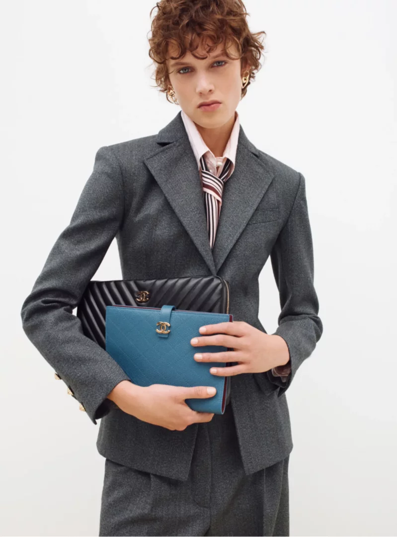 Kısa kıvırcık saçlı, gri Chanel blazer ceket, pembe gömlek ve çizgili kravat giyen bir model, elinde siyah ve turkuaz renkli, ikonik CC logolu Chanel çantalar tutarak doğrudan izleyiciye bakıyor.
