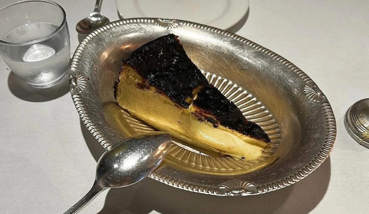 Kremamsı dokusu ve davetkar görünümüyle dikkat çeken, pürüzsüz yüzeyi ve hafifçe kızarmış kenarlarıyla beyaz bir tabakta sunulan klasik bir cheesecake dilimi, tatlı severler için vazgeçilmez bir lezzet deneyimi sunuyor.