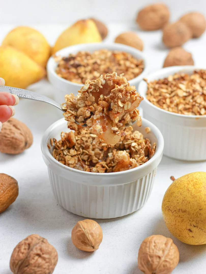 Bir kaşık dolusu yulaflı armut crumble, beyaz bir fırın kabından kaldırılırken yakın çekimde görülüyor; arka planda bulanık duran diğer crumble kapları ve etrafa dağılmış taze armutlar ile cevizler, bu lezzetli tatlının doğal ve besleyici içeriğini vurguluyor.