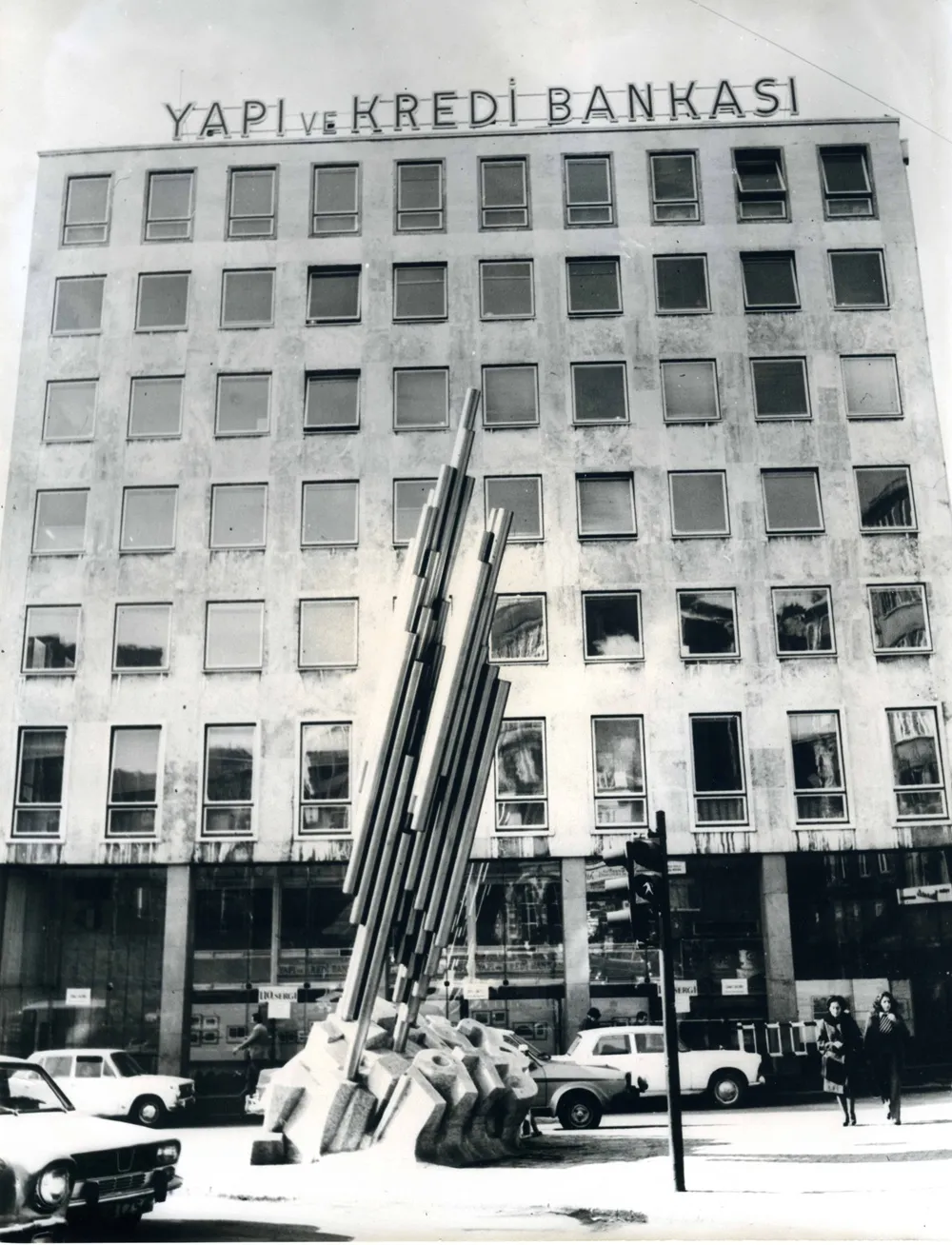Siyah beyaz fotoğrafta, Sadi Çalık'ın modern ve soyut Cumhuriyet 50. Yılı Anıtı, üzerinde YAPI VE KREDİ BANKASI yazan yüksek bir binanın önünde, eski model arabaların ve birkaç yayanın bulunduğu bir şehir caddesinde yer alıyor.