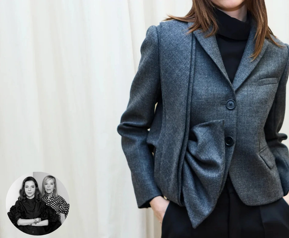 Siyah boğazlı bir kazak ve pantolon üzerine, ön kısmında heykelsi ve özgün kıvrım detaylarına sahip antrasit gri bir blazer ceket giyen, elleri ceplerinde duran bir kadını gösteren bu görselin sol alt köşesinde, Dice Kayek markasının yaratıcı tasarımcılarının siyah beyaz bir portresi yuvarlak bir çerçevede yer alıyor.