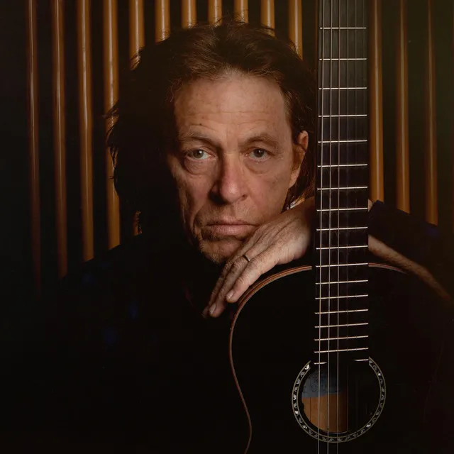 Gitarist ve besteci Dominic Miller, düşünceli ifadesi ve hafif dağınık saçlarıyla doğrudan objektife bakarken, sağ elini koyu renkli klasik gitarının gövdesine dayamış, arkasında sıcak tonlarda dikey ahşap panellerin yer aldığı bu yakın plan fotoğrafıyla solo kariyerinin olgunluğunu sergiliyor.