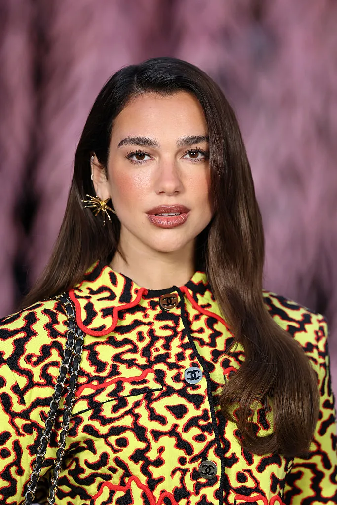 Koyu renk saçları geriye taranmış, belirgin göz makyajı ve sade ama şık, açık renk bir bluz giyen Dua Lipa'nın kendine özgü ciddi ve kararlı ifadesiyle objektife odaklandığı, arka planın bulanık olduğu profesyonel bir çekimden yayımlanan etkileyici portresi.