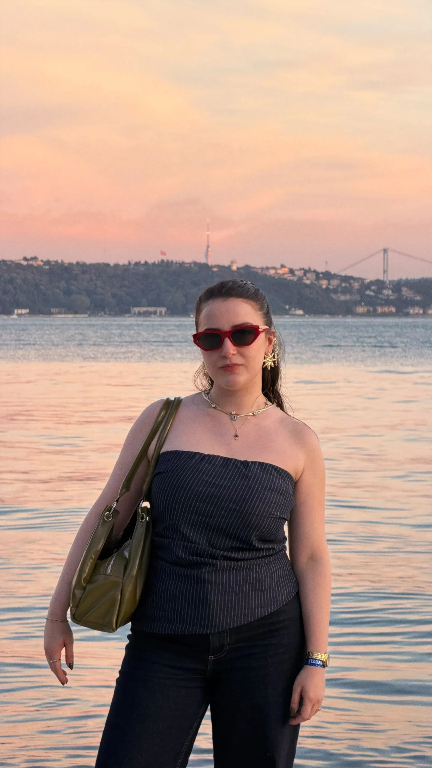 Kırmızı kedi gözü güneş gözlüğü, yıldız ve sallantılı küpeler, straplez çizgili bir üst, koyu renk kot pantolon ve haki yeşili bir omuz çantasıyla poz veren genç bir kadın, gün batımının pembe ve turuncu yansımalarıyla parlayan denizin kenarında duruyor; arka planda ağaçlarla kaplı şehir silüeti, Çamlıca Kulesi'ne benzeyen bir yapı ve köprü silüeti görünüyor.