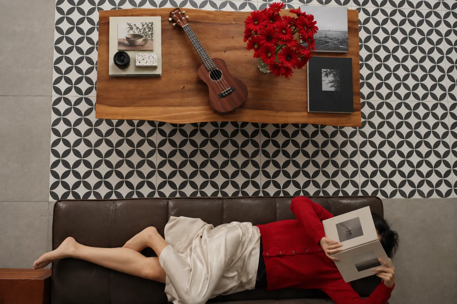 Siyah ve beyaz geometrik desenli zemine sahip bir odada, kahverengi bir kanepede uzanmış, yüzünü okuduğu kitaba gizlemiş bir kişi, doğal ahşap sehpasında duran ukulele ve kırmızı çiçeklerle çevrili, evde kaliteli zaman geçirerek slow living felsefesiyle iyi yaşamın tadını çıkarıyor.