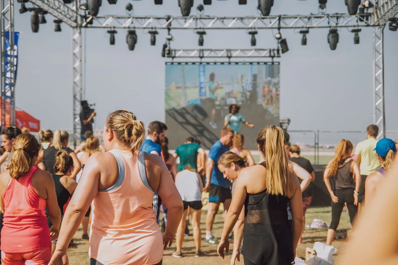 Güneşli bir günde, arkadan görülen kalabalık bir grup insan, sahnede büyük bir ekranda gösterilen bir eğitmen eşliğinde açık havada bir fitness etkinliğine katılırken, bu canlı sahne bir fitness tatili deneyimini akla getiriyor.