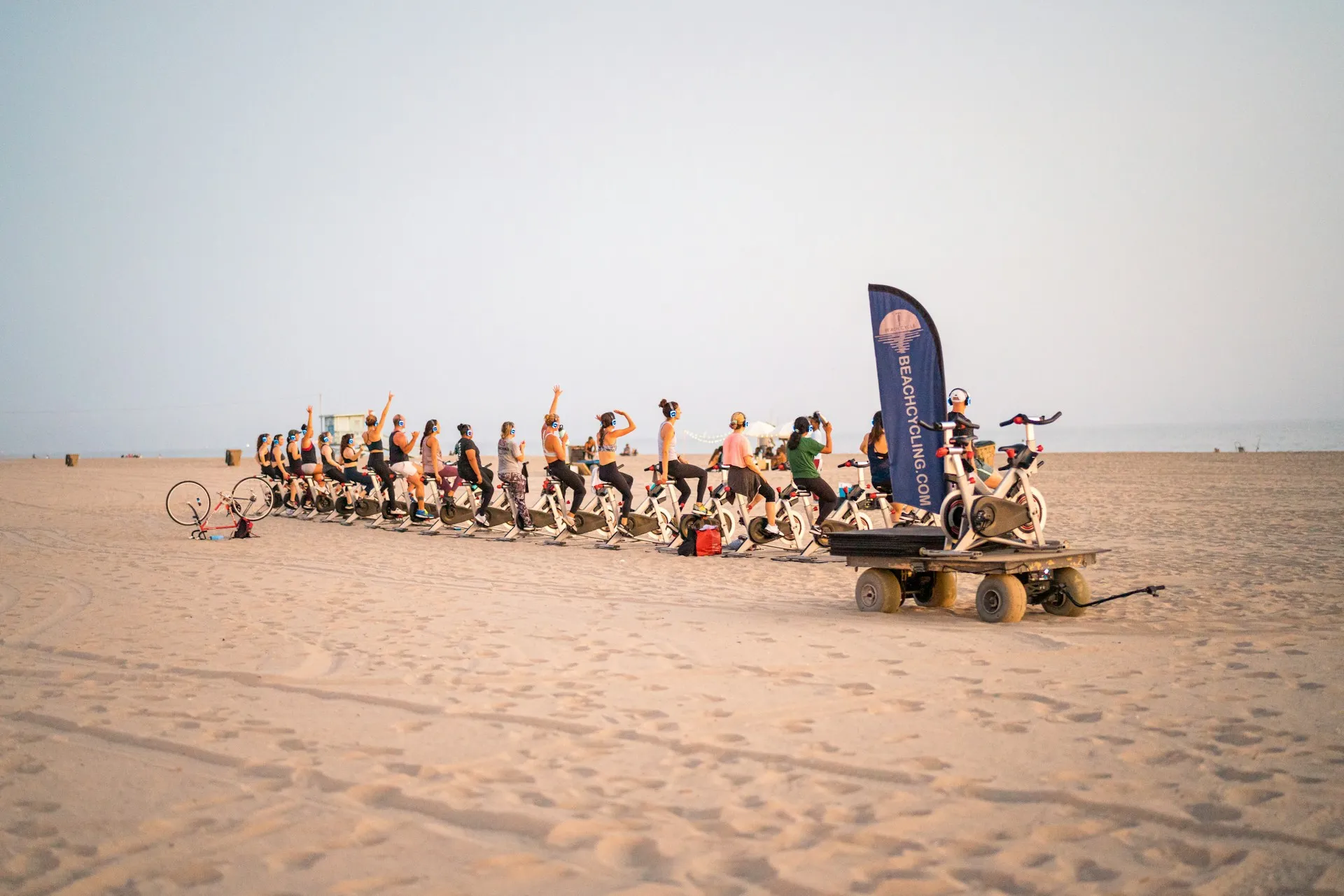 Geniş kumsalda, Beachcycling.com pankartı önünde kulaklık takmış çok sayıda kişinin spin bisikletlerinde coşkuyla pedallayarak açık havada bir fitness aktivitesine katıldığı, fitness turizminin dinamizmini yansıtan keyifli bir an.