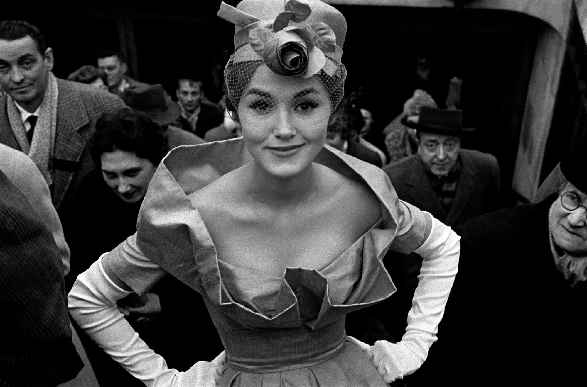 Frank Horvat'ın moda fotoğrafını stüdyodan sokağın gerçekliğine taşıyan tarzını yansıtan bu fotoğrafta, koyu renkli palto ve zarif şapka giyen bir kadın, kalabalık bir şehir sokağında doğal ve anlık bir ifadeyle yürürken objektife yansıyor.