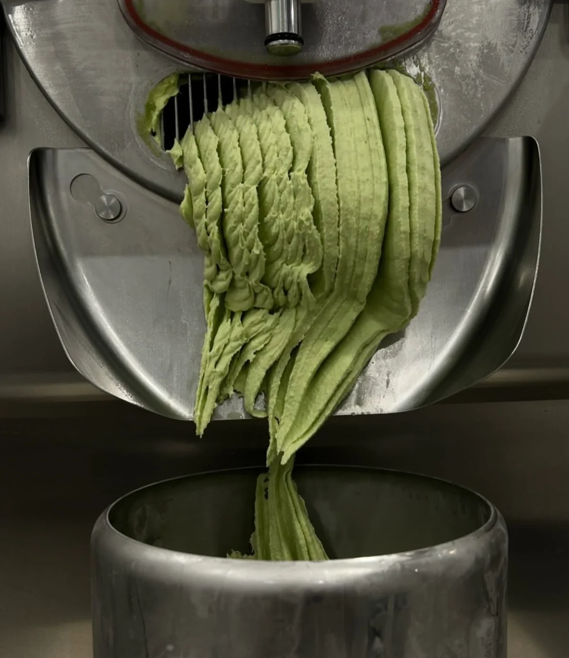 Paslanmaz çelik bir makineden, parlak yeşil ve kıvrımlı dokuya sahip taze yapılmış gelato'nun büyük bir metal kaba akarak doluşunu gösteren bu yakın çekim, ürünün üretim aşamasını detaylı bir şekilde sunuyor.