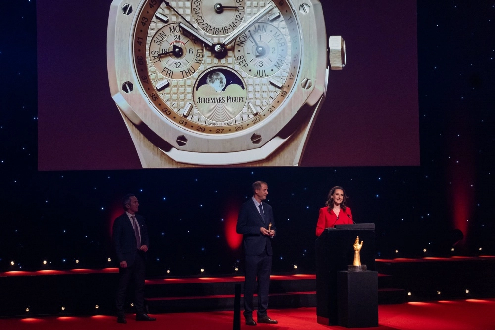 Kırmızı halı kaplı, ışıklı bir sahnede, büyük bir ekranda Audemars Piguet lüks saatinin detaylı görüntüsü sergilenirken, GPHG ödül töreninde bir kadın kürsüde konuşuyor, iki erkek de küçük bir ödülle sahnede yer alıyor.