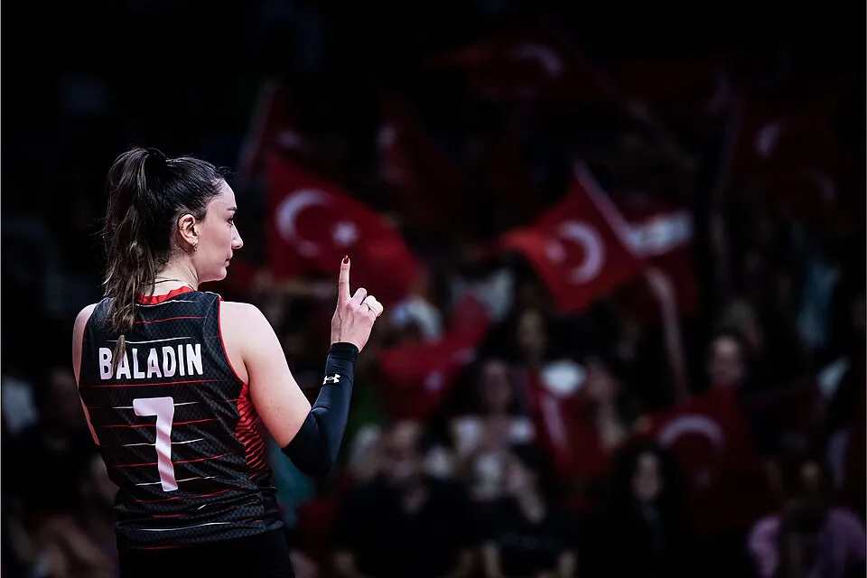 Hande Baladın'ın sırtı dönük ve hafif yandan göründüğü bu fotoğrafta, üzerinde ismi ve 7 numarası yazan siyah formasını giymiş voleybolcu, sağ işaret parmağını yukarı kaldırmış durumda, arka planda ise Filenin Sultanları'nı destekleyen Türk bayraklarıyla coşkulu bir kalabalık bulanık bir şekilde görülüyor.