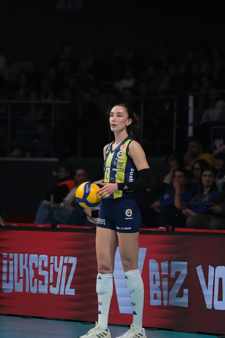 Fenerbahçe'nin 7 numaralı lacivert formasıyla voleybol sahasında sarı-mavi bir topu tutan, izleyicilerle dolu tribünler önünde kariyerindeki azmini yansıtan kararlı ifadesiyle duran voleybolcu Hande Baladın.