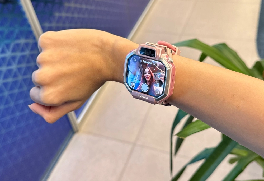 Bir kişinin bileğinde takılı olan pembe ve gümüş detaylı Huawei akıllı çocuk saati, geniş ekranında canlı kamera görüntüsüyle genç bir kadının gülümseyen selfisini ve fotoğraf çekme arayüzünü net bir şekilde gösteriyor.